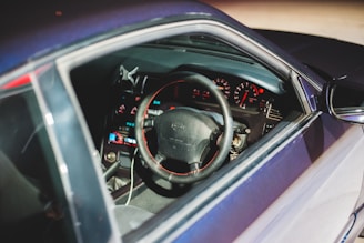 A car interior view focusing on the steering wheel and dashboard. The dashboard displays illuminated gauges and indicators, with a brightly lit digital interface. The steering wheel features a black finish with red stitching.
