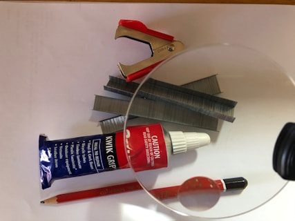 A collection of stationary and office items includes a red and silver staple remover, stacks of metallic staples, a blue tube of adhesive labeled 'KWIK GRIP', a bottle with a red caution label, a red pencil, and a black pen. These items are arranged on a white surface and partially under a magnifying glass that distorts a part of the pencil.