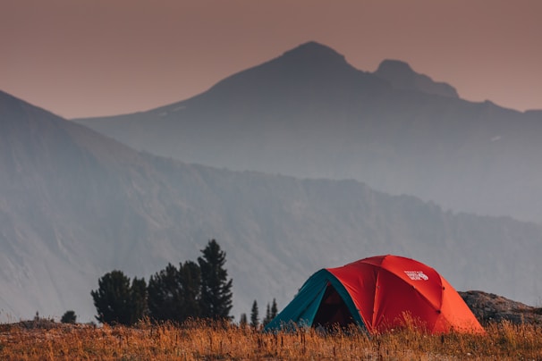 A vibrant red and blue tent is set up on a grassy field. In the background, there are layered mountains fading into the distance under a hazy sky. Trees are visible nearby, enhancing the secluded and natural setting.