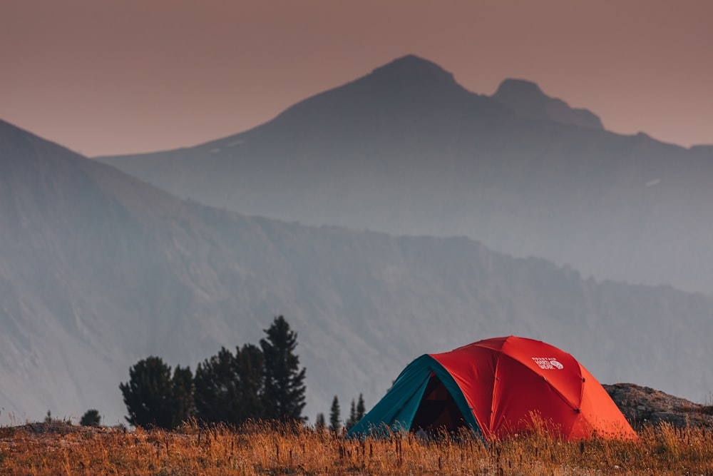 tenda rossa sul campo di erba marrone vicino agli alberi verdi e alla montagna durante il giorno