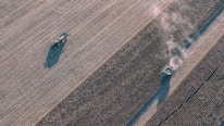 An aerial view of a large agricultural field with a tractor pulling equipment and a harvester emitting dust as it moves along rows of crops. The field is characterized by its neat, parallel lines created by the rows of crops.