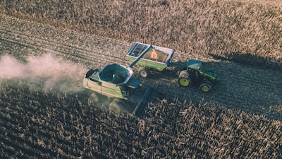 Aerial view of a combine harvester operating in a vast field, collecting crops. A tractor with an attached trailer follows behind, filled with harvested produce. Dust trails form in the wake of the machinery, creating a dynamic scene illustrating agricultural work.