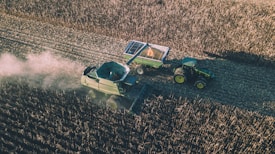 Aerial view of a combine harvester operating in a vast field, collecting crops. A tractor with an attached trailer follows behind, filled with harvested produce. Dust trails form in the wake of the machinery, creating a dynamic scene illustrating agricultural work.