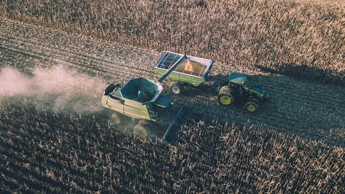 Aerial view of a combine harvester operating in a vast field, collecting crops. A tractor with an attached trailer follows behind, filled with harvested produce. Dust trails form in the wake of the machinery, creating a dynamic scene illustrating agricultural work.