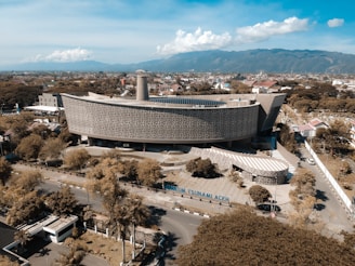 An aerial view of a large, modern architectural building surrounded by trees and roads. The structure, identified as Museum Tsunami Aceh, is characterized by its unique, wave-like design with numerous perforations on its facade. Mountains and a partly cloudy sky serve as the backdrop, with a sprawling cityscape visible in the background.