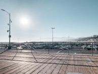 A wooden deck with metal railings overlooks a large parking lot filled with cars. The sun is high in the clear sky, casting long shadows on the deck. Distant hills are faintly visible along with a few buildings on the right side. The ocean appears to be in the far background.