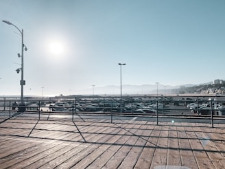 A wooden deck with metal railings overlooks a large parking lot filled with cars. The sun is high in the clear sky, casting long shadows on the deck. Distant hills are faintly visible along with a few buildings on the right side. The ocean appears to be in the far background.