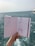 A hand holds a sketchbook open to reveal fashion illustrations against a background of the sea. The sketches depict clothing designs, including a long dress and a jumpsuit, both enhanced with detailed patterns. The ocean is visible in the distance, with slight waves under a muted sky.