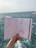 A hand holds a sketchbook open to reveal fashion illustrations against a background of the sea. The sketches depict clothing designs, including a long dress and a jumpsuit, both enhanced with detailed patterns. The ocean is visible in the distance, with slight waves under a muted sky.