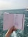 A hand holds a sketchbook open to reveal fashion illustrations against a background of the sea. The sketches depict clothing designs, including a long dress and a jumpsuit, both enhanced with detailed patterns. The ocean is visible in the distance, with slight waves under a muted sky.