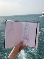 A hand holds a sketchbook open to reveal fashion illustrations against a background of the sea. The sketches depict clothing designs, including a long dress and a jumpsuit, both enhanced with detailed patterns. The ocean is visible in the distance, with slight waves under a muted sky.