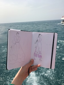 A hand holds a sketchbook open to reveal fashion illustrations against a background of the sea. The sketches depict clothing designs, including a long dress and a jumpsuit, both enhanced with detailed patterns. The ocean is visible in the distance, with slight waves under a muted sky.