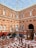 A spacious courtyard with a glass ceiling and red brick walls featuring arched windows. Rows of empty red-cushioned chairs are arranged on a patterned floor. There is an ornate wooden organ and a setup for a musical performance, including a harp and a grand piano. Several people are scattered around, appearing to be setting up or preparing for an event.