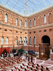 A spacious courtyard with a glass ceiling and red brick walls featuring arched windows. Rows of empty red-cushioned chairs are arranged on a patterned floor. There is an ornate wooden organ and a setup for a musical performance, including a harp and a grand piano. Several people are scattered around, appearing to be setting up or preparing for an event.