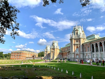 A grand and historic palace with intricately designed architecture featuring tall towers, arches, and ornate details. It is set in a lush green park with a clear blue sky above, scattered with fluffy white clouds. Visitors are walking around the grounds enjoying the scenery and taking photos.