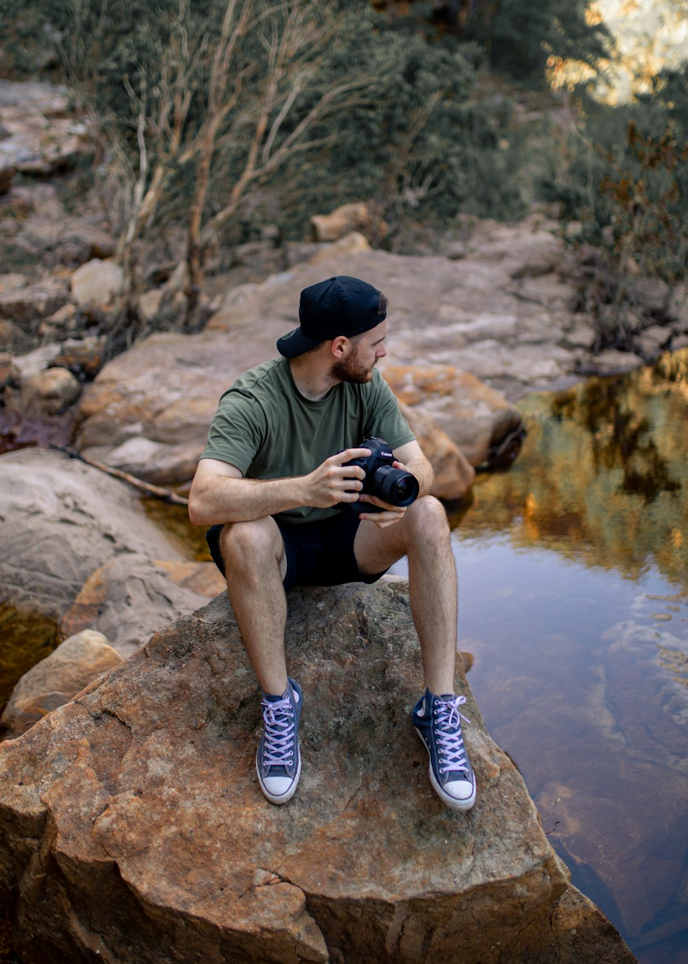 man in green t-shirt and brown shorts holding black dslr camera sitting on brown rock