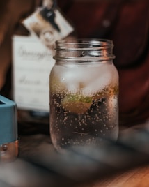 A close-up of a mason jar filled with a fizzy beverage containing ice and a lime slice. The background includes a blurred image of a bottle with a label, giving a cozy and rustic feel.
