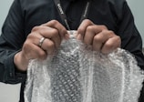 Hands are gripping a sheet of bubble wrap, with a person wearing a black shirt and a lanyard visible. A ring is worn on the left hand, and the bubble wrap is being pulled taut, suggesting potential for popping.