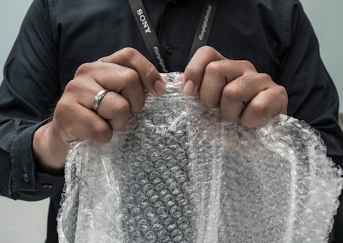 Hands are gripping a sheet of bubble wrap, with a person wearing a black shirt and a lanyard visible. A ring is worn on the left hand, and the bubble wrap is being pulled taut, suggesting potential for popping.