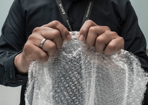 Hands are gripping a sheet of bubble wrap, with a person wearing a black shirt and a lanyard visible. A ring is worn on the left hand, and the bubble wrap is being pulled taut, suggesting potential for popping.