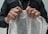 Hands are gripping a sheet of bubble wrap, with a person wearing a black shirt and a lanyard visible. A ring is worn on the left hand, and the bubble wrap is being pulled taut, suggesting potential for popping.
