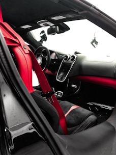 The interior of a luxury car features a sleek design with red and black leather seats and a red seat belt. The dashboard is modern, with a central touch screen and several sophisticated controls. The steering wheel appears sporty, and the rearview mirror is visible alongside the side mirror. The overall design emphasizes elegance and performance.