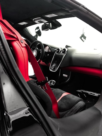 The interior of a luxury car features a sleek design with red and black leather seats and a red seat belt. The dashboard is modern, with a central touch screen and several sophisticated controls. The steering wheel appears sporty, and the rearview mirror is visible alongside the side mirror. The overall design emphasizes elegance and performance.