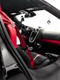 The interior of a luxury car features a sleek design with red and black leather seats and a red seat belt. The dashboard is modern, with a central touch screen and several sophisticated controls. The steering wheel appears sporty, and the rearview mirror is visible alongside the side mirror. The overall design emphasizes elegance and performance.