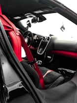 The interior of a luxury car features a sleek design with red and black leather seats and a red seat belt. The dashboard is modern, with a central touch screen and several sophisticated controls. The steering wheel appears sporty, and the rearview mirror is visible alongside the side mirror. The overall design emphasizes elegance and performance.