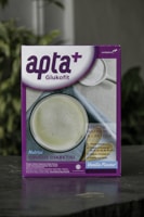 A box featuring a health-related product labeled 'apta+ Glukofit' with a purple and white design. The product claims a vanilla flavor and highlights benefits for diabetes, including low sodium and lactose content, high fiber, vitamins, and calcium. The front design includes a photo of a white beverage to suggest consumption.