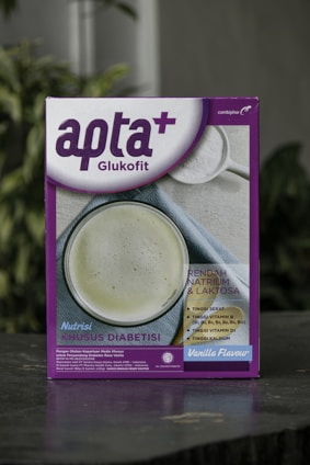 A box featuring a health-related product labeled 'apta+ Glukofit' with a purple and white design. The product claims a vanilla flavor and highlights benefits for diabetes, including low sodium and lactose content, high fiber, vitamins, and calcium. The front design includes a photo of a white beverage to suggest consumption.