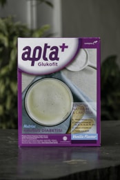 A box featuring a health-related product labeled 'apta+ Glukofit' with a purple and white design. The product claims a vanilla flavor and highlights benefits for diabetes, including low sodium and lactose content, high fiber, vitamins, and calcium. The front design includes a photo of a white beverage to suggest consumption.