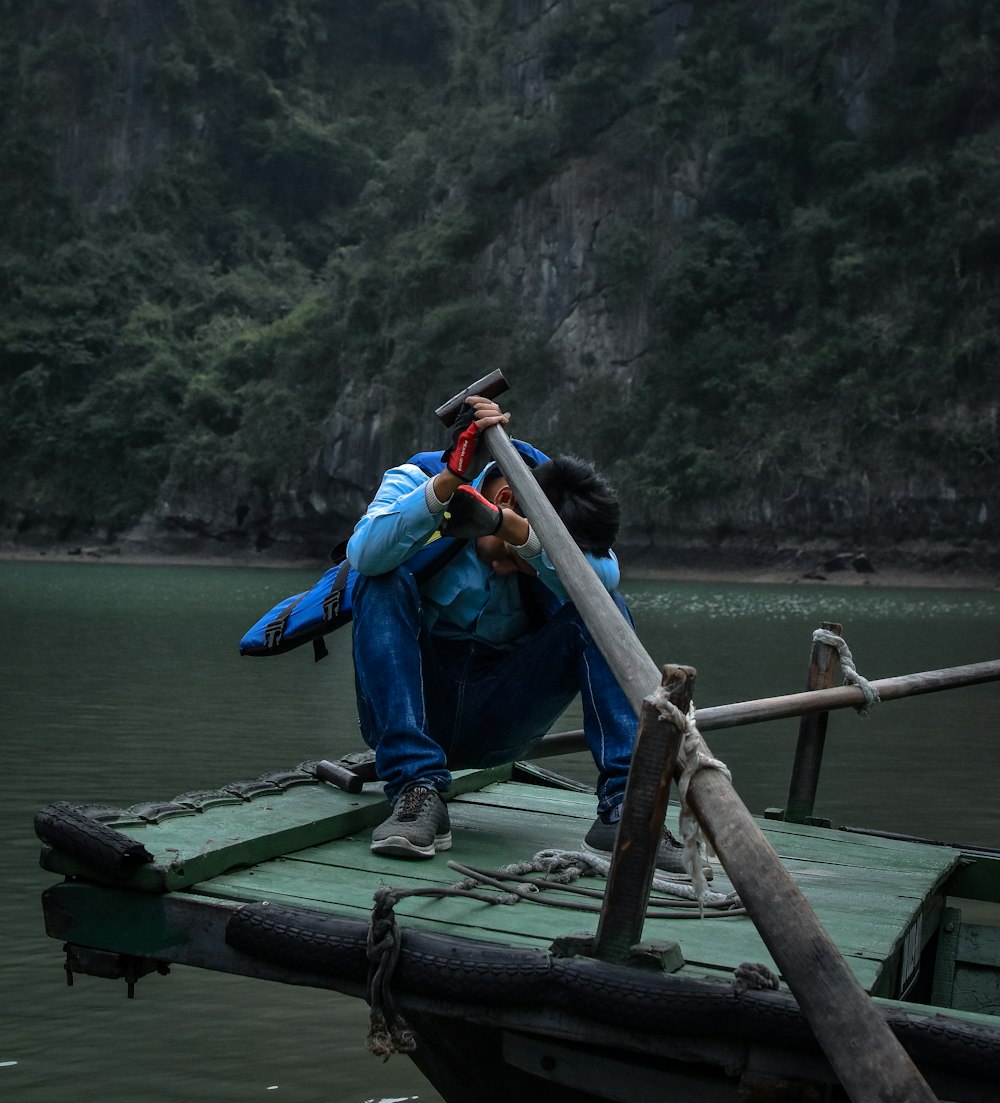 man in blue jacket and blue denim jeans sitting on brown wooden dock during daytime