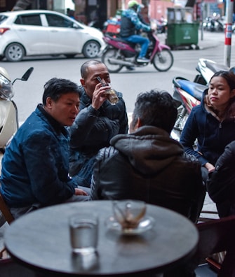 A group of people sitting close together at a small table, engaging in conversation. One person is drinking from a glass while others listen and interact. The setting appears to be an outdoor or street-side café with motorcycles and cars parked nearby.