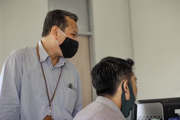Two individuals wearing face masks are situated in an office environment, with one standing and observing a computer screen that the other is using.