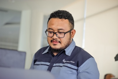 A man with glasses is focused on his work in a modern office setting, wearing a dark uniform. The background is softly blurred, emphasizing the clean and professional environment.