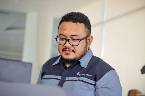 A man with glasses is focused on his work in a modern office setting, wearing a dark uniform. The background is softly blurred, emphasizing the clean and professional environment.