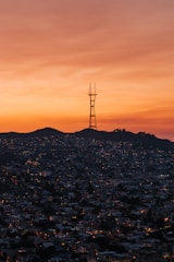 A cityscape at dusk with a prominent radio tower situated on a hill. The sky exhibits vibrant shades of orange and red, casting a warm glow over the densely packed buildings below. Small lights from buildings illuminate the city, creating a lively and bustling atmosphere.