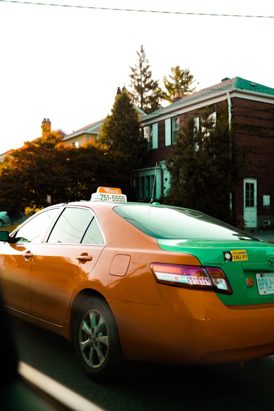 A green and orange taxi with a visible phone number on its sign is parked on a street in front of residential houses. The surrounding area includes trees and a typical suburban neighborhood environment.
