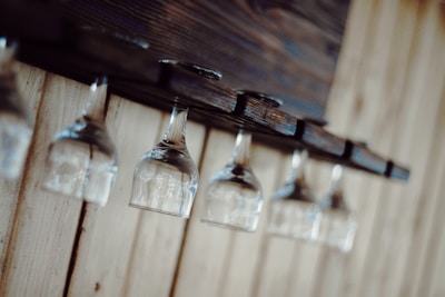 A series of upside-down wine glasses hang from a wooden rack against a light wooden panel wall, creating a rustic and organized aesthetic.