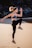 A person is performing a high-intensity exercise, executing a jump with one knee lifted and arms in motion. The setting is an outdoor sandy beach with rocky formations in the background. The person is wearing athletic gear including a sports bra, leggings, and running shoes.