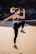 A person is performing a high-intensity exercise, executing a jump with one knee lifted and arms in motion. The setting is an outdoor sandy beach with rocky formations in the background. The person is wearing athletic gear including a sports bra, leggings, and running shoes.