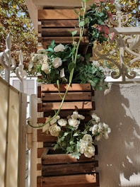 Wooden planters are mounted vertically on a wall, filled with lush green foliage and white flowers. The setup is sunlit, casting intricate shadows against the wall and a wrought iron fence. Some light pink flowers peep at the top, adding a touch of color. The overall arrangement has an elegant and natural aesthetic.