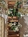 Wooden planters are mounted vertically on a wall, filled with lush green foliage and white flowers. The setup is sunlit, casting intricate shadows against the wall and a wrought iron fence. Some light pink flowers peep at the top, adding a touch of color. The overall arrangement has an elegant and natural aesthetic.