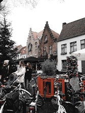 A European street scene showcases charming brick and white buildings adorned with festive decorations and wreaths. The cobblestone street is bustling with people, including women and children, some with bicycles and strollers. One of the buildings features a distinctive stepped gable design. Christmas trees and holiday lights add to the cheerful atmosphere.