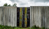 A weathered, corrugated metal fence with a central gate made of vertically placed wooden planks, some of which have chipped paint showing layers of green, blue, and yellow. The fence stands amidst a grassy area with some visible small plants and bushes, while the cloudy sky looms overhead.