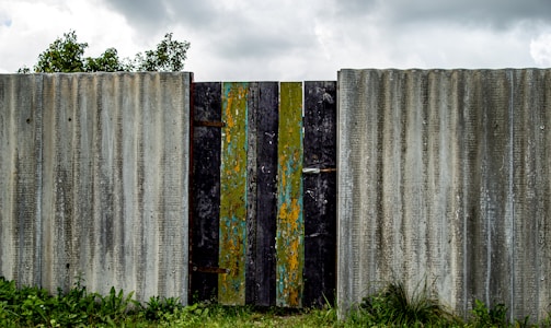 A weathered, corrugated metal fence with a central gate made of vertically placed wooden planks, some of which have chipped paint showing layers of green, blue, and yellow. The fence stands amidst a grassy area with some visible small plants and bushes, while the cloudy sky looms overhead.