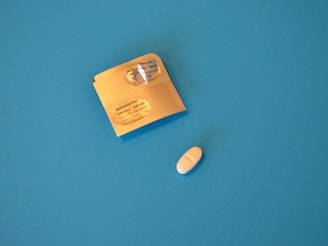 A single white pill is placed next to a partially opened blister pack on a solid blue background. The packaging is golden yellow with text indicating it is Azithromycin, a 500 mg film-coated tablet.