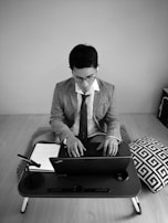 A person wearing a suit and tie sits cross-legged at a small desk, working on a laptop. A notebook with handwritten notes and a microphone are placed next to the laptop. The setup is on a wooden floor with a geometric-patterned cushion beside it.