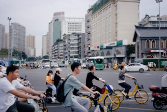 People are riding bicycles and scooters in a busy urban intersection surrounded by tall buildings. Cars and a bus are also visible in the background, along with several pedestrians.